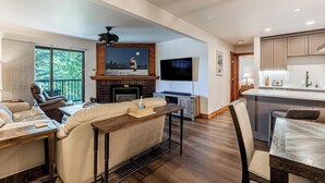 Ein modernes Wohnzimmer mit Kamin, einem an der Wand befestigten Flachbildfernseher, einem Essbereich mit Holztisch und einer Küche mit Arbeitsplatte und Schränken.