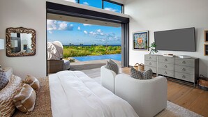A modern bedroom with a large bed, a sofa, a TV, and a view of a pool and greenery. - Hale Mahina Hou: Luxe 4BR Home w/ Ocean Views, Infinity Pool & Outdoor Seating (Koloa)