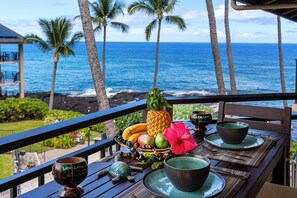 A wooden table with a tropical fruit arrangement, a hibiscus flower, and two bowls on a balcony overlooking the ocean with palm trees. - Kona Makai 6303: Tropical Retreat 1BR w/ Ocean Views, Pool & Gym (Kailua Kona)