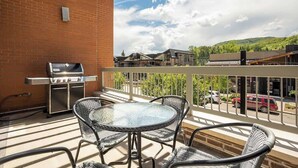 A balcony with a grill, wicker chairs, and a view of buildings and trees. - Sprawling Condo | Central Downtown | Free Shuttle (Steamboat Springs)