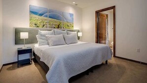 A hotel room with a bed, a nightstand, a wall-mounted lamp, and a door leading to another room. - Sprawling Condo | Central Downtown | Free Shuttle (Steamboat Springs)