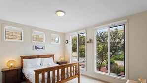 A bedroom with a wooden bed, bedside lamps, a wall clock, and large windows with a view of greenery.