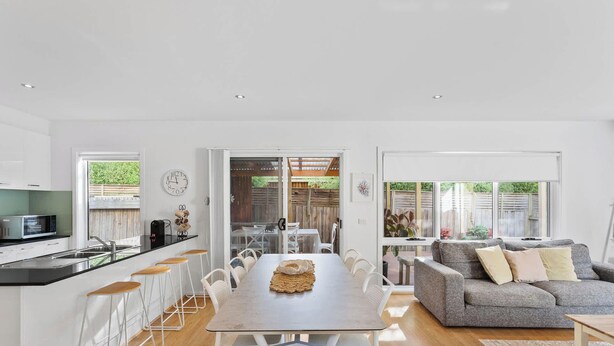 A modern kitchen with a microwave, bar stools, and a dining table with chairs. There is a grey sofa and a window with a view of greenery.