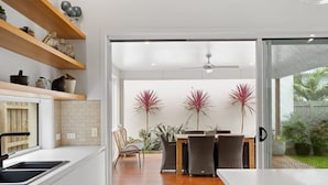 A modern kitchen with wooden shelves, a dining area with wicker chairs, and a view of a garden through glass doors. - Family Luxury 3 Bdr King Bed. (Newhaven)