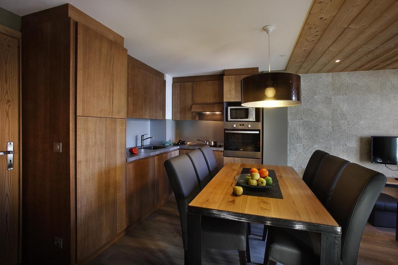 A modern kitchen with wooden cabinets, a dining table with chairs, and a fruit bowl.