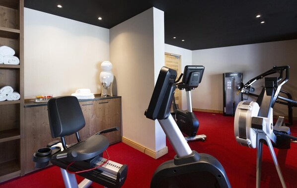 Une salle de sport équipée de machines de cardio, un meuble en bois contenant des serviettes et un sol recouvert d’une moquette rouge.