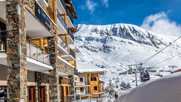 A multi-story building with stone and wood exterior, a ski lift in the background, and a snow-covered mountain. - Wifi, 150m from ski slopes, fitness, swimming pool, sauna, hammam, balcony, 98m², Alpe d'Huez (L'Alpe d'Huez)