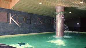 Una piscina interior con una gran fuente vertical en el centro, rodeada por un suelo de baldosas oscuras y una pared con el nombre 'Kohinoor' en letras grandes.