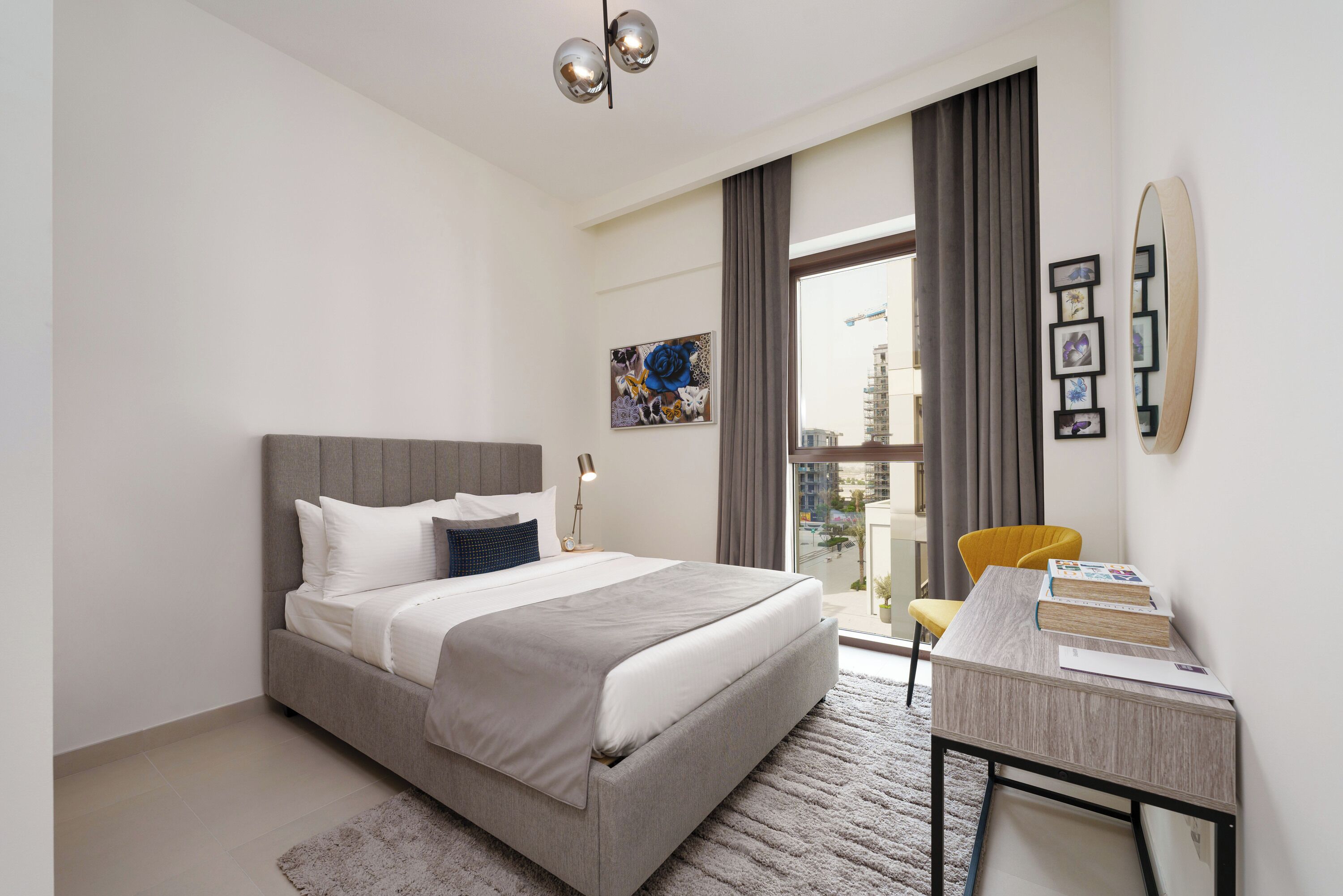 A modern hotel room with a large bed, a desk with books, a city view, and a painting on the wall.