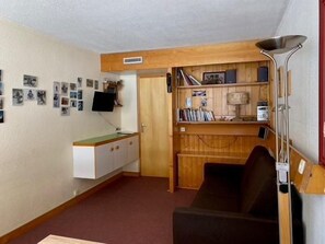 A compact living space with a kitchenette, a sofa, a TV mounted on the wall, and a bookshelf.