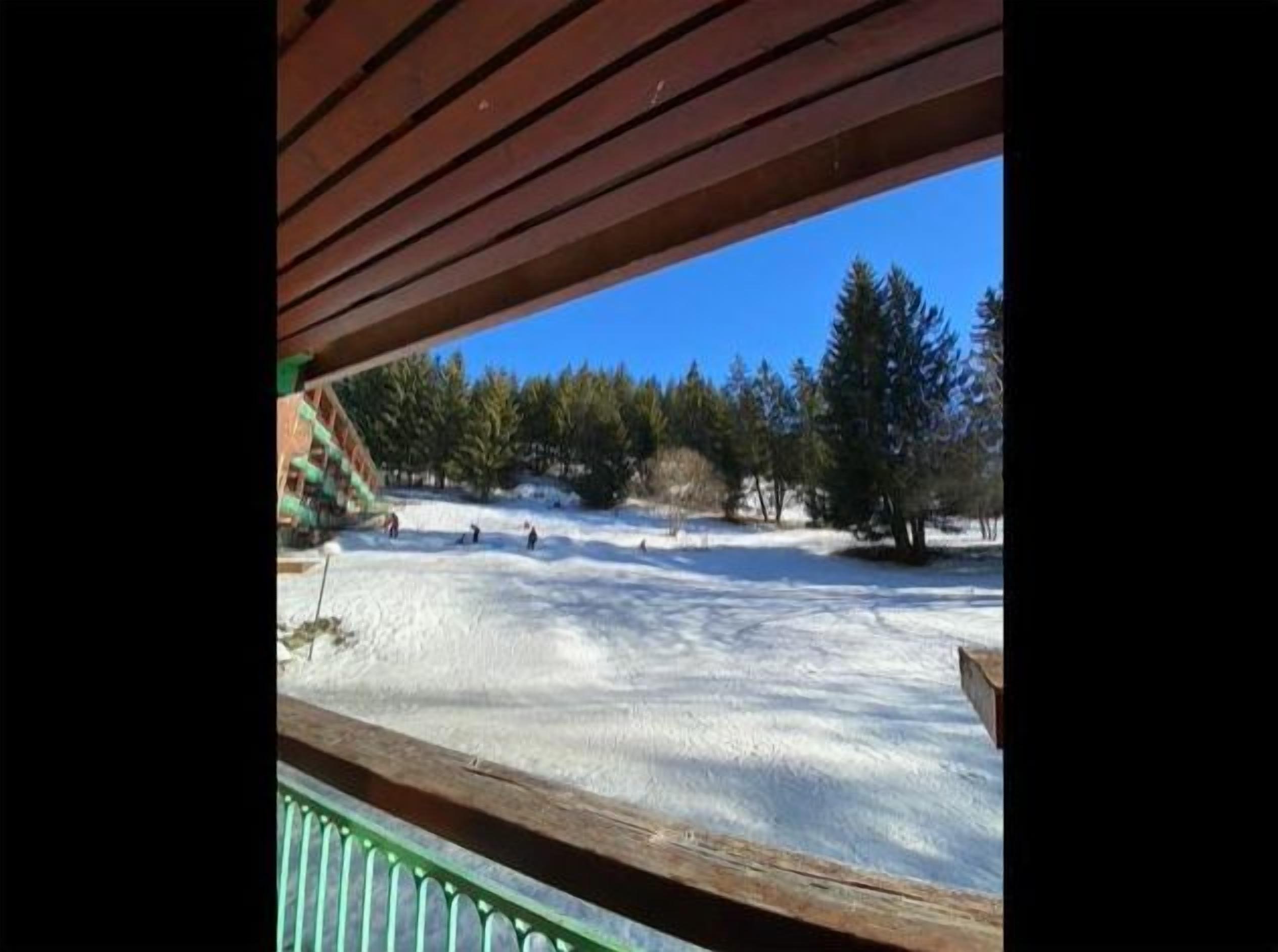 A snowy outdoor area with a wooden deck and a view of trees and a clear sky.