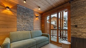A cozy living room with a sofa, stone wall, wooden ceiling, and a view of a wooden cabin through large glass doors.