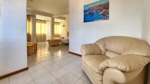 A living room with a beige sofa, a painting of a coastal scene, and a view of a bedroom with a bed and curtains. - Txada Hostel (Praia)