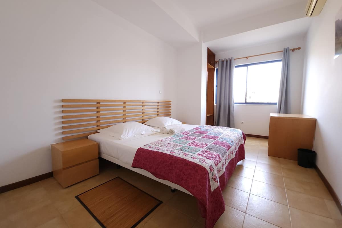 A bedroom with a wooden bed frame, a red and white quilt, a wooden nightstand, a small wooden chest, and a window with curtains.