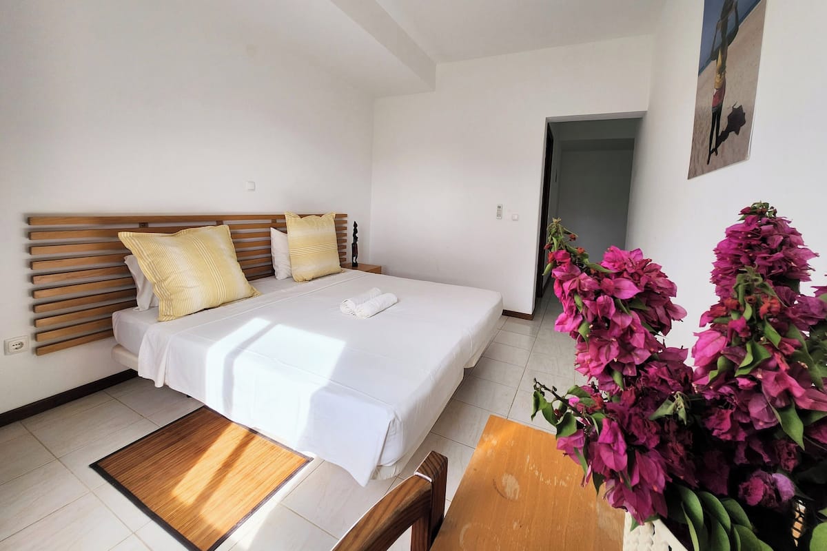 A bedroom with a bed, wooden headboard, white bedding, a small wooden table, and a chair.