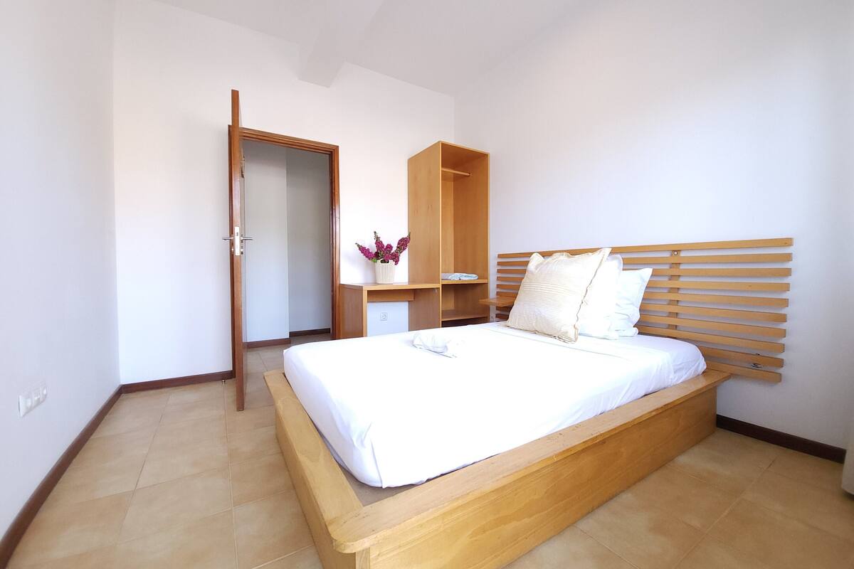 A bedroom with a wooden bed, a wooden wardrobe, a small bedside table, and a door leading to another room.