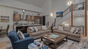 A modern living room with a sofa, coffee table, and chairs. There is a kitchen area with cabinets and a dining table. A staircase leads to an upper level.