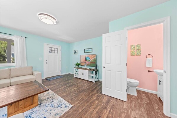 Una habitación con un sofá color beige, una mesa de centro de madera, una puerta blanca, una pared rosa, un inodoro blanco y un pequeño armario con una planta.