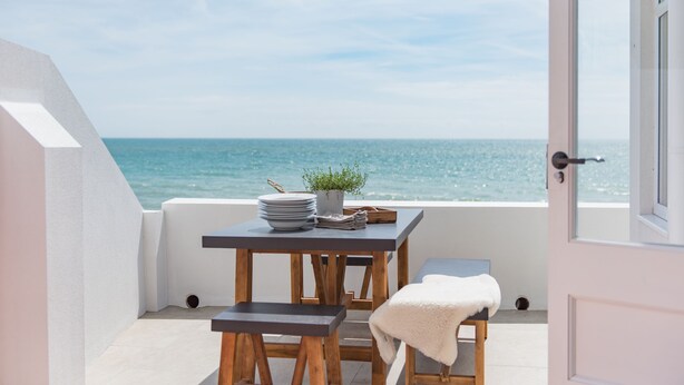 Uma varanda com vista para o mar, uma mesa com pratos e uma planta, e uma porta.