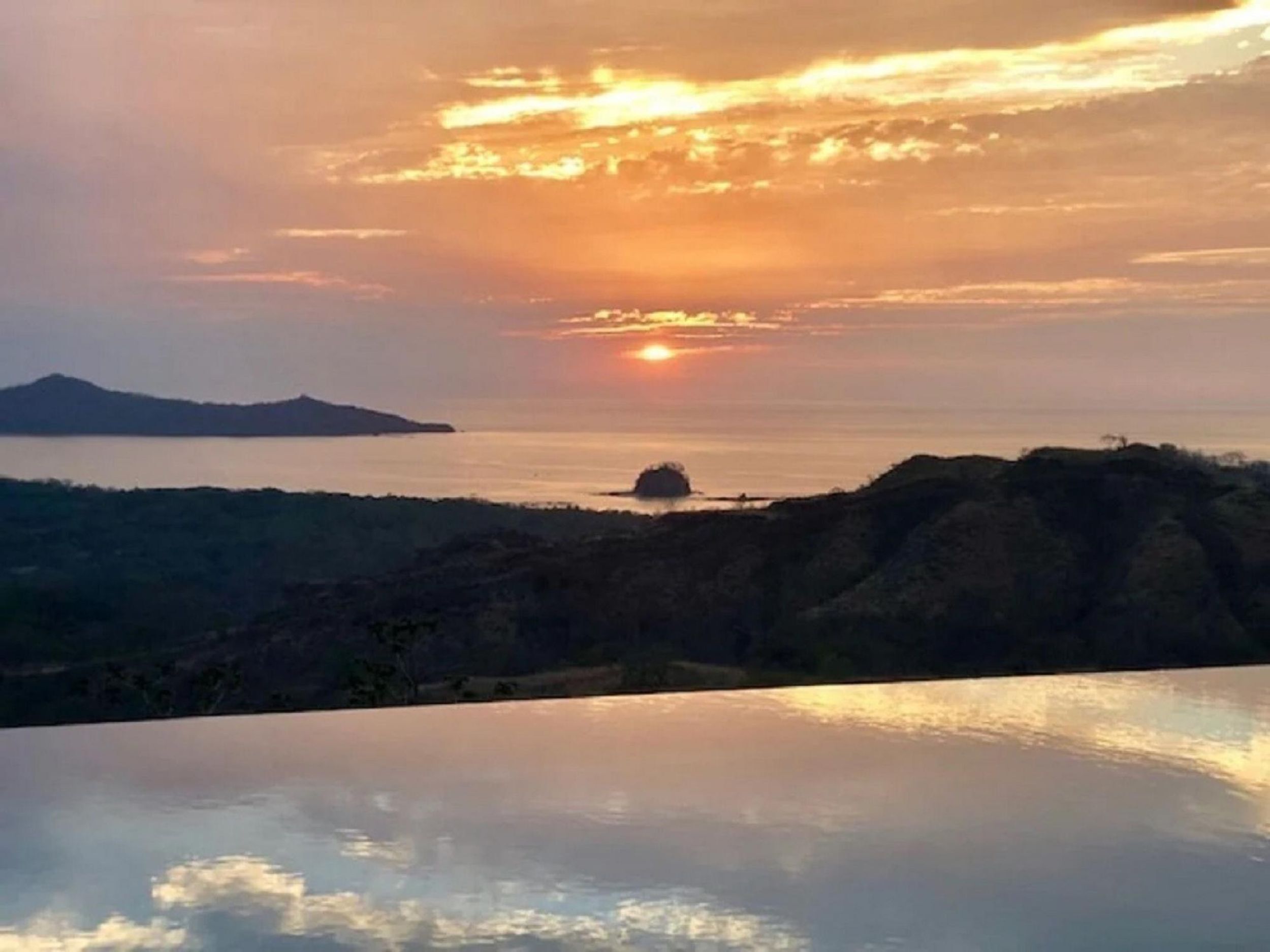 A sunset over a body of water with a distant landmass and a rocky outcrop.