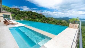 Una piscina infinita rectangular con agua azul cristalina, rodeada por una terraza blanca y sillones de madera.