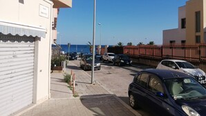 Une rue résidentielle bordée de voitures garées, un ciel dégagé et une vue sur la mer en arrière-plan.
