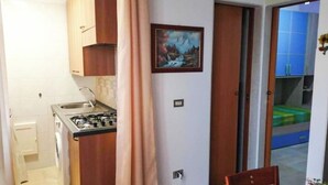 Une cuisine compacte équipée d’une cuisinière à gaz, d’un évier et de meubles de rangement en bois. Un tableau orne le mur et une porte donne accès à un balcon.