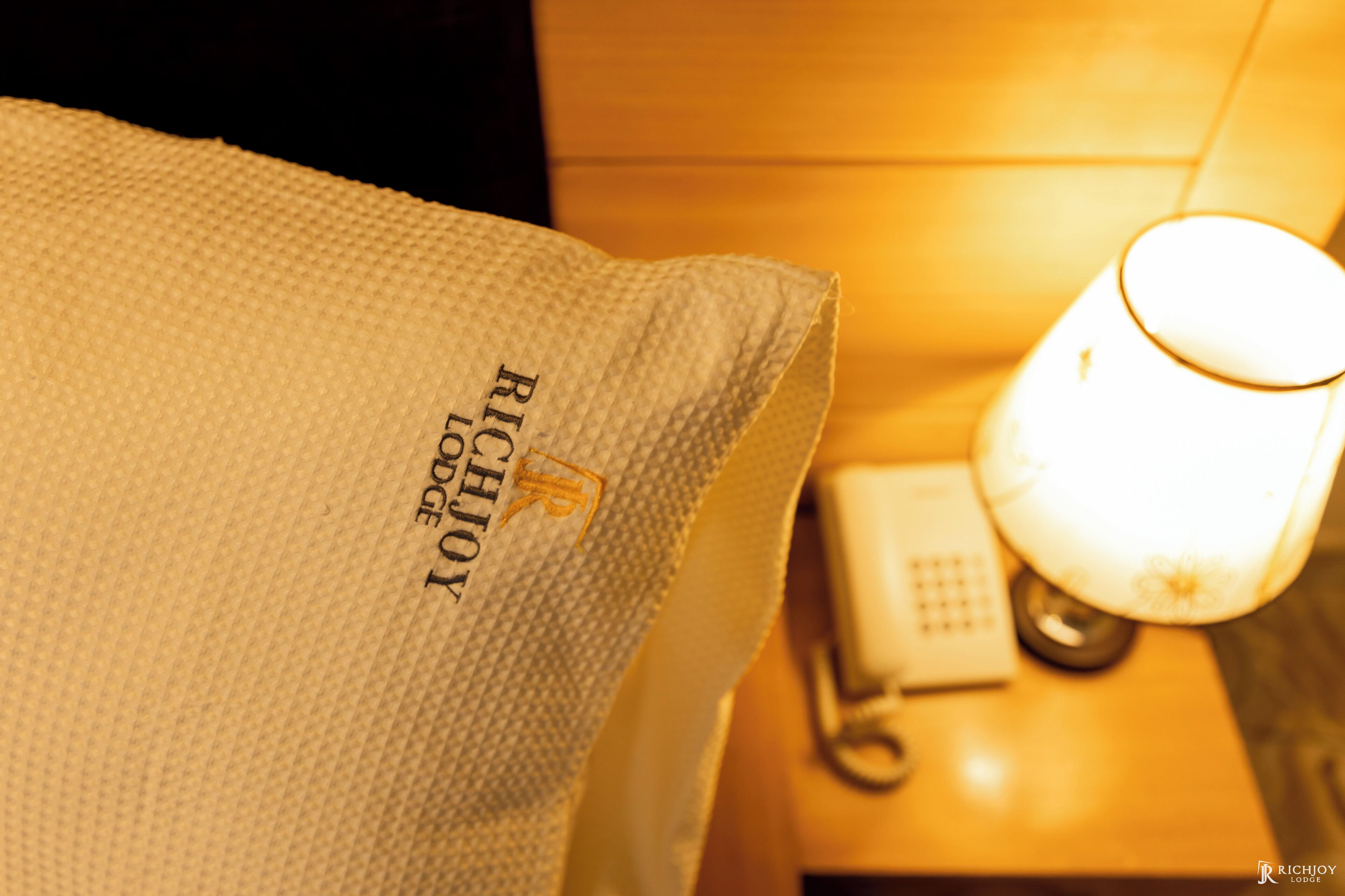 Un oreiller d’hôtel orné du mot « RichJoy » brodé dessus, une lampe de chevet et un téléphone posés sur une table de chevet en bois.