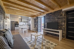 A modern living room with a stone wall, wooden ceiling beams, a flat-screen TV, and a dining area with white furniture. - Mountain Retreat by Homie (Porto Moniz)