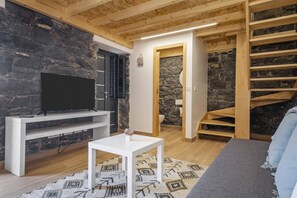 A modern living room with a stone wall, a flat-screen TV on a white stand, a small white table, and a staircase with wooden steps. - Mountain Retreat by Homie (Porto Moniz)