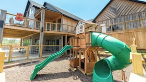 A wooden play structure with a green slide and a playhouse, set against a backdrop of a large wooden building with balconies.