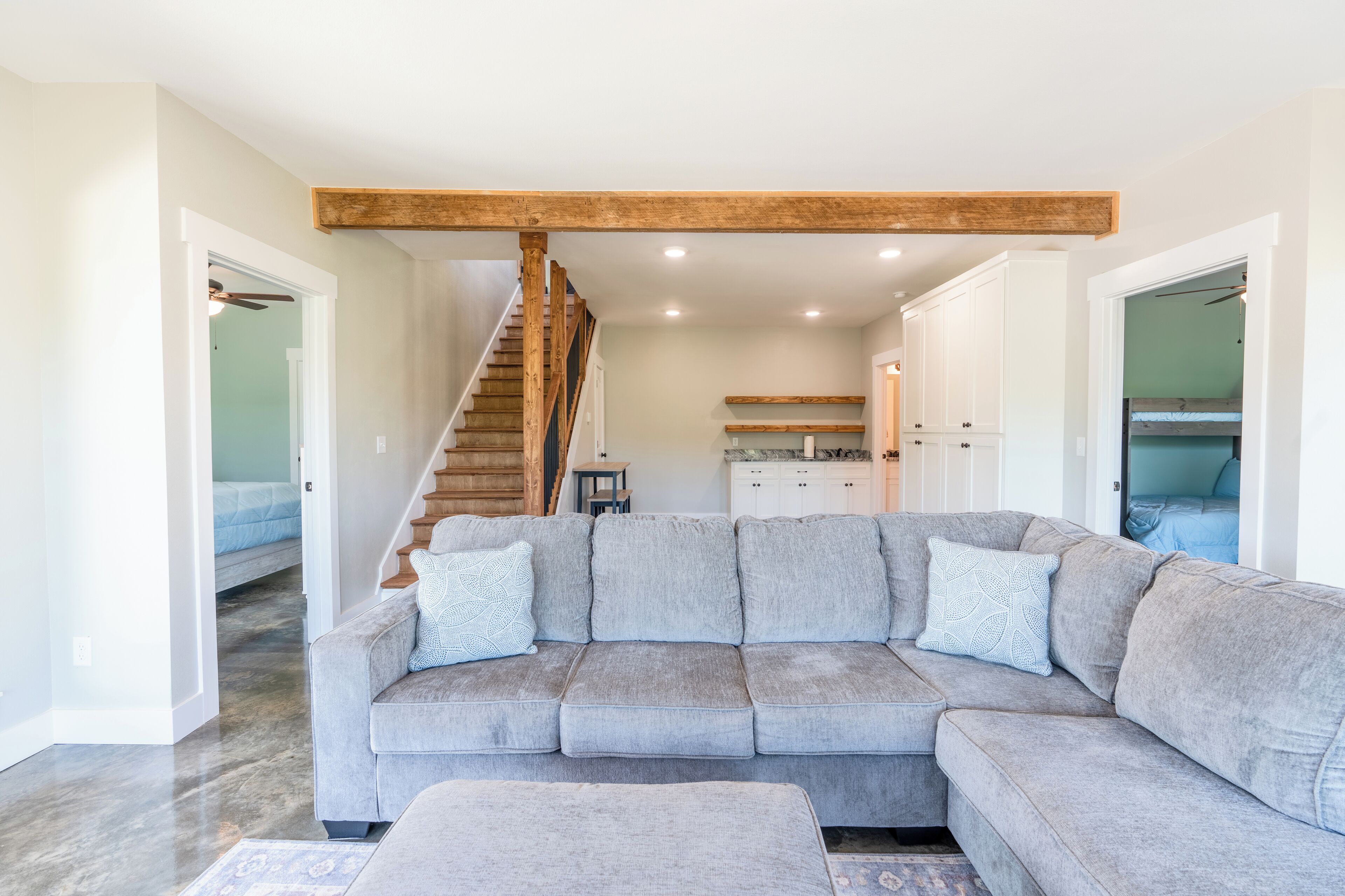 A modern living room with a grey sectional sofa, wooden beams above, a staircase, and a kitchen in the background.
