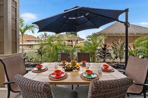Een eettafel buiten opstelling met rieten stoelen, een grote parasol en een tropische achtergrond.