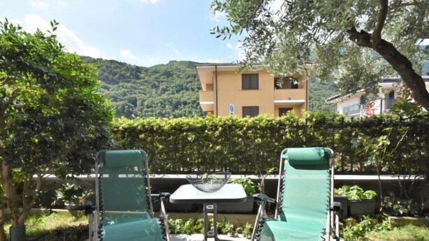 Two green lounge chairs with metal frames and a small table in between, set on a patio with a view of a yellow building and green hills in the background.