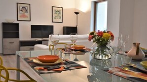 A modern living room with a glass dining table set for two, a sofa, a TV, and artwork on the walls.