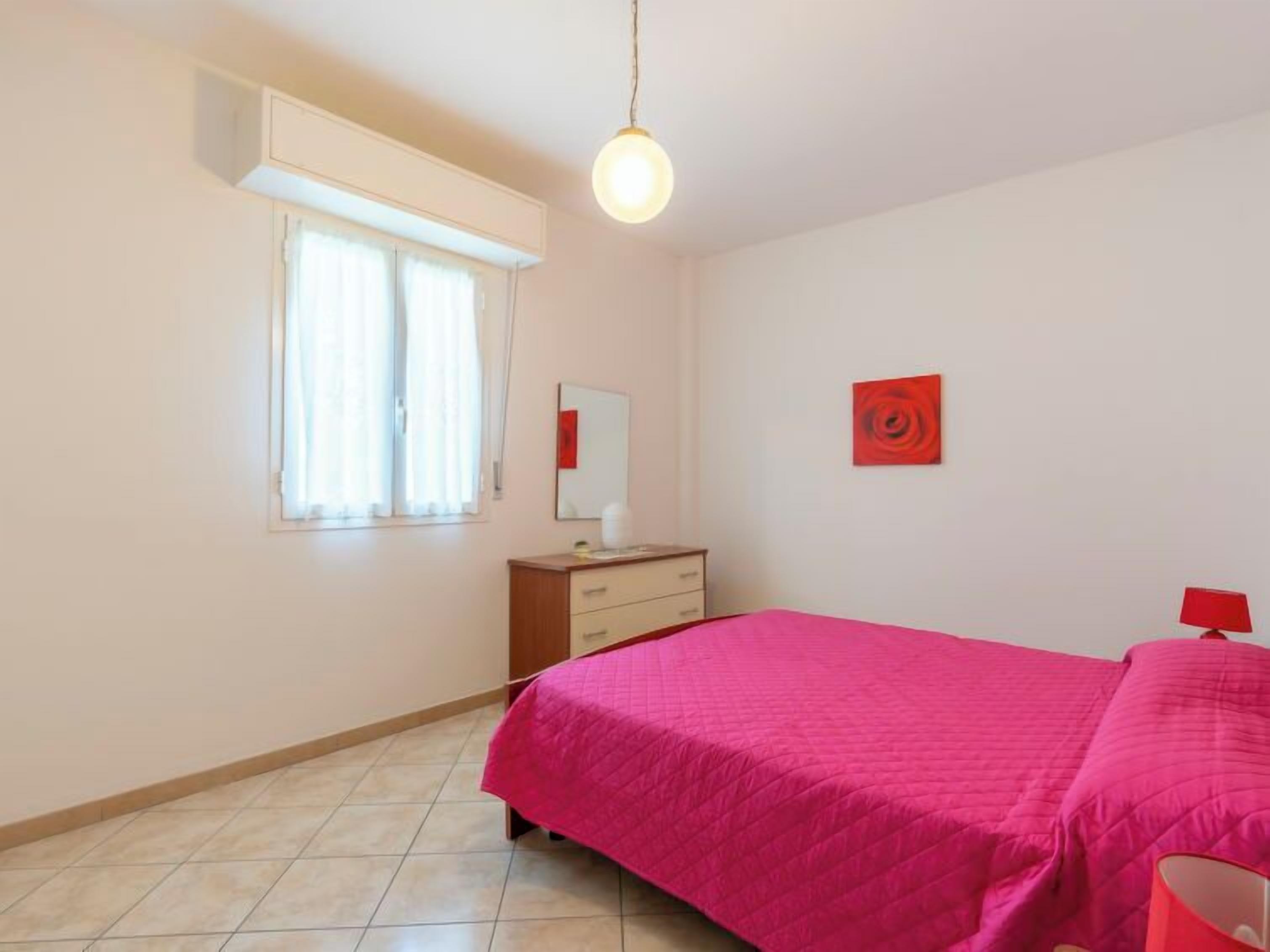 A bedroom with a bed, a dresser, a window with curtains, a wall-mounted mirror, and a red artwork.