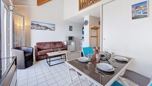 A dining area with a table set for four, a sofa, a coffee table, and a view of a balcony with a wooden railing. - Parc de Pontaillac by Interhome (Vaux Sur Mer)