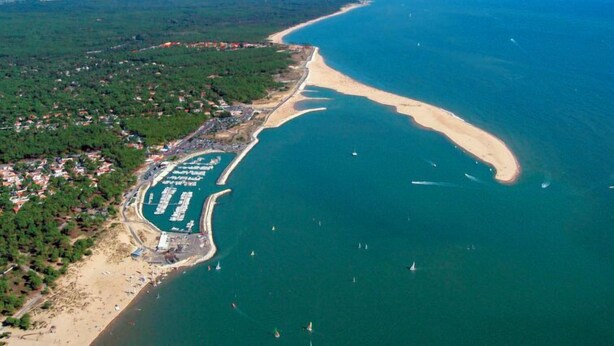 Vue aérienne d’une zone côtière comprenant un port de plaisance, une plage de sable et une végétation luxuriante environnante.