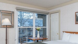 Une chambre d’hôtel avec un lit, une fenêtre donnant sur des arbres, une table avec un vase de fleurs et une lampe.