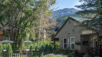 A mountain lodge with a welcoming sign, surrounded by lush greenery and a view of mountains.