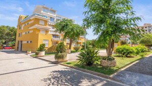 Um edifício residencial amarelo de vários andares, com varandas, rodeado por vegetação bem cuidada e um céu limpo.