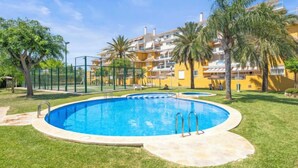 Piscina com cerca ao redor, palmeiras e um edifício amarelo com varandas ao fundo.