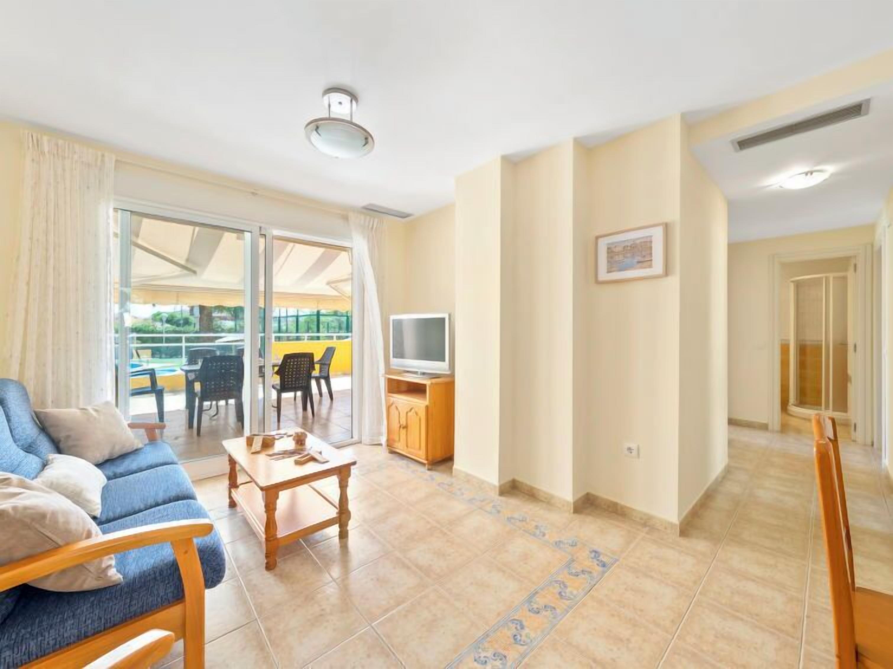 Ein modernes Wohnzimmer mit Sofa, Couchtisch und Fernseher. Ein Balkon mit Gartenmöbeln ist vorhanden.