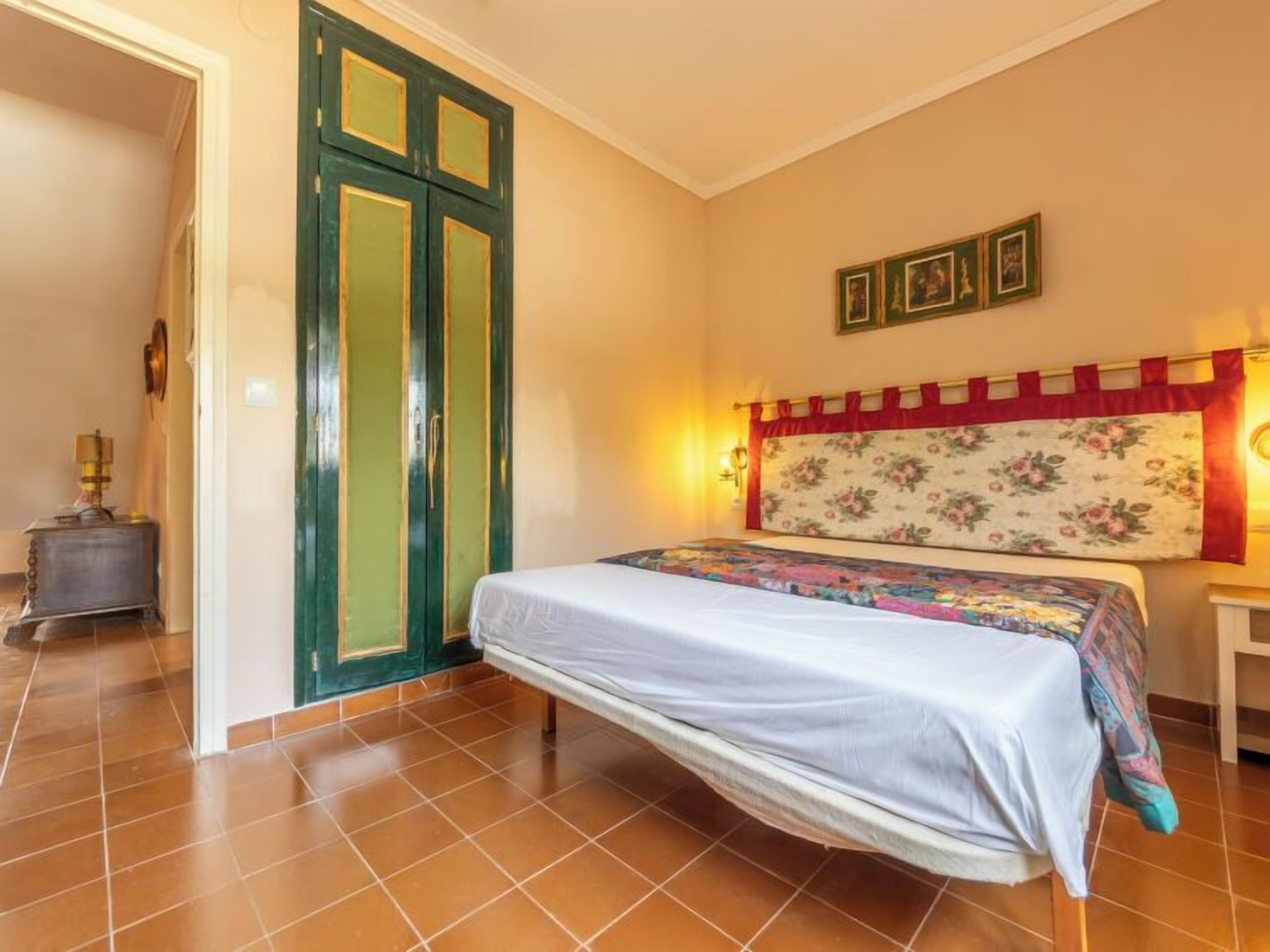 A bedroom with a bed, a nightstand, a lamp, a door, and a wall with framed pictures.