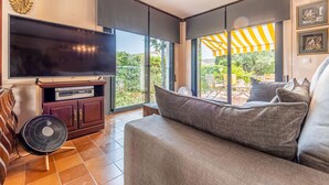 Un salon équipé d’un téléviseur à écran plat, d’un meuble en bois, d’un ventilateur et d’une grande fenêtre donnant sur un paysage verdoyant.