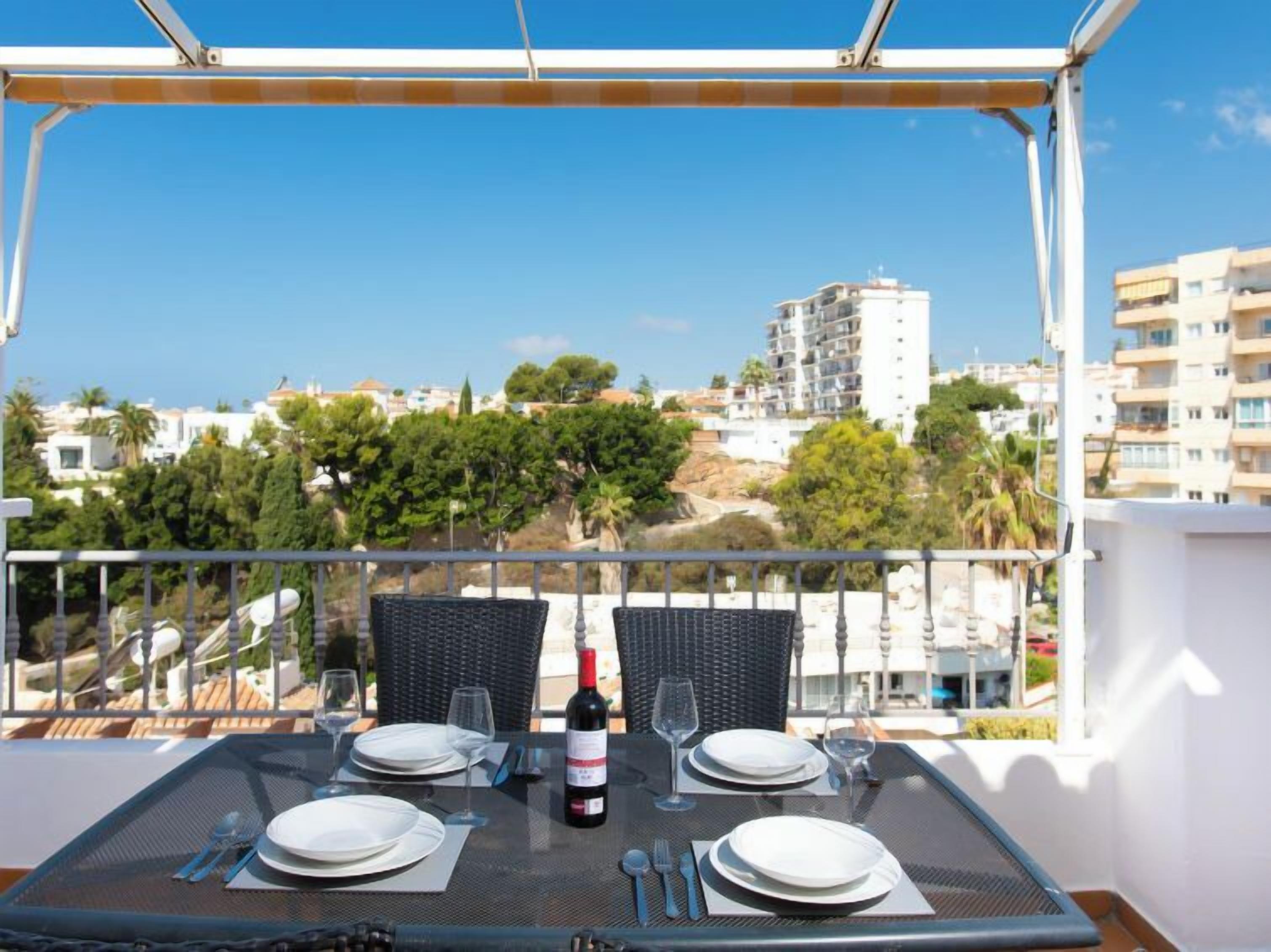 A balcony with a table set for two, overlooking a cityscape with buildings and trees.