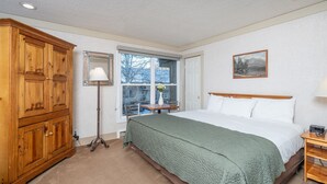 A bedroom with a bed, a wooden wardrobe, a floor lamp, a window with a view, and a painting on the wall.