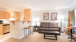 A modern living room with a sofa, coffee table, and a dining area with a table and chairs. There is a kitchen area with wooden cabinets and a dishwasher. - Mountainside Inn 106 1 Bedroom Condo by Alpine Lodging Telluride (Telluride)