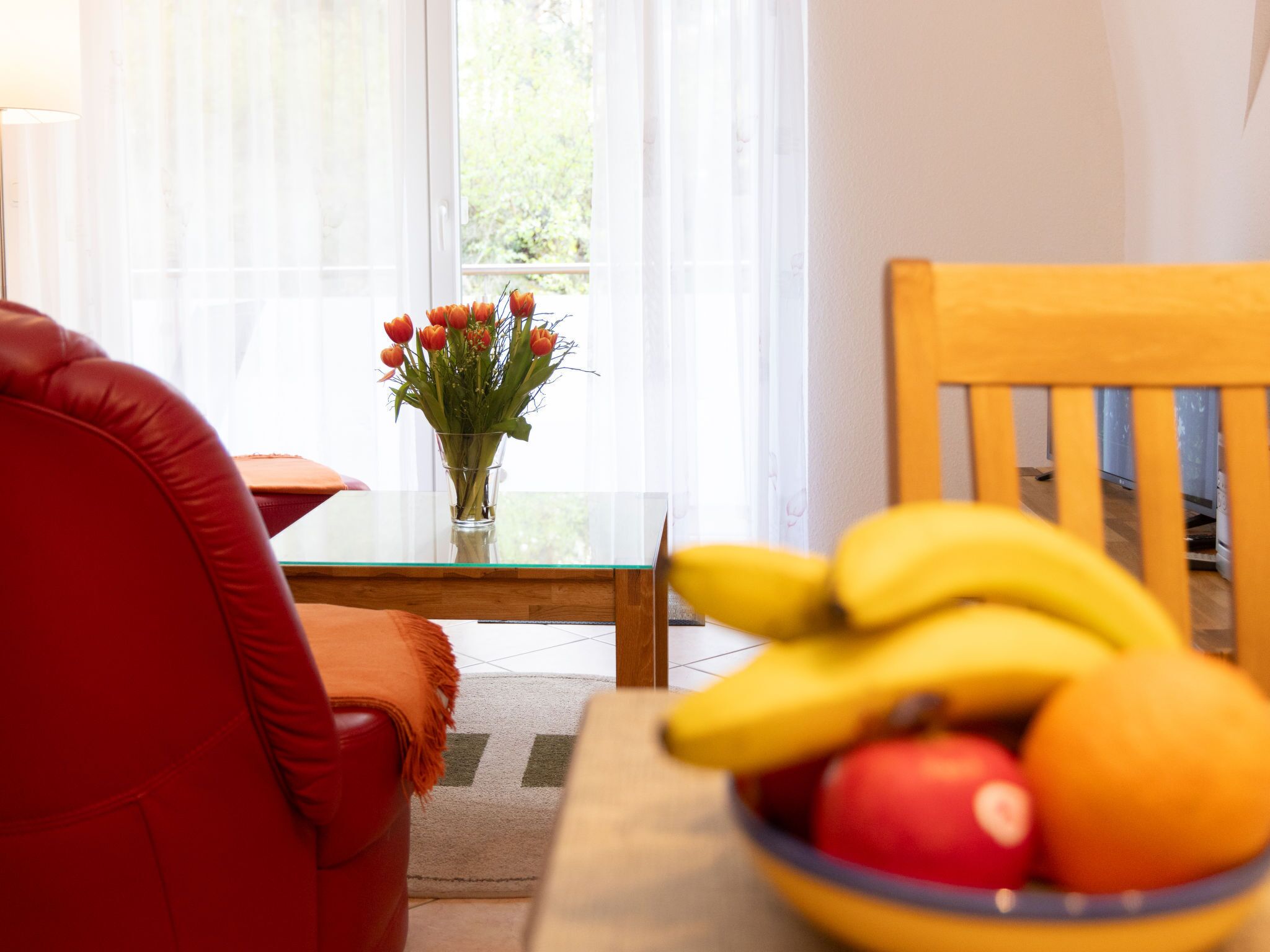 Ein Wohnzimmer mit Glastisch, Blumenvase, rotem Sessel und Obstschale.