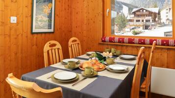 Un coin repas avec une table dressée pour quatre personnes, des chaises en bois, un tableau encadré au mur et une fenêtre donnant sur un paysage de montagne et des bâtiments.